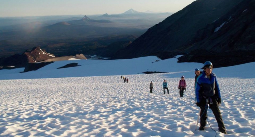 A group of outward bound students wearing mountaineering gear make their way up a snowy slope. There are mountains in the distance. 